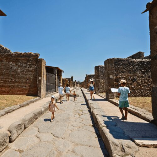 ourists, including families with children, exploring the ancient stone-paved streets of Pompeii under a bright blue sky.